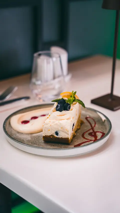 A slice of cheesecake with blueberries and a raspberry drizzle at an evening table