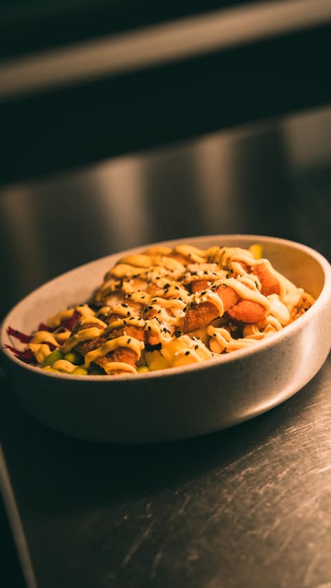 A poke bowl topped with crispy breaded pieces, sesame seeds and a drizzle of sauce